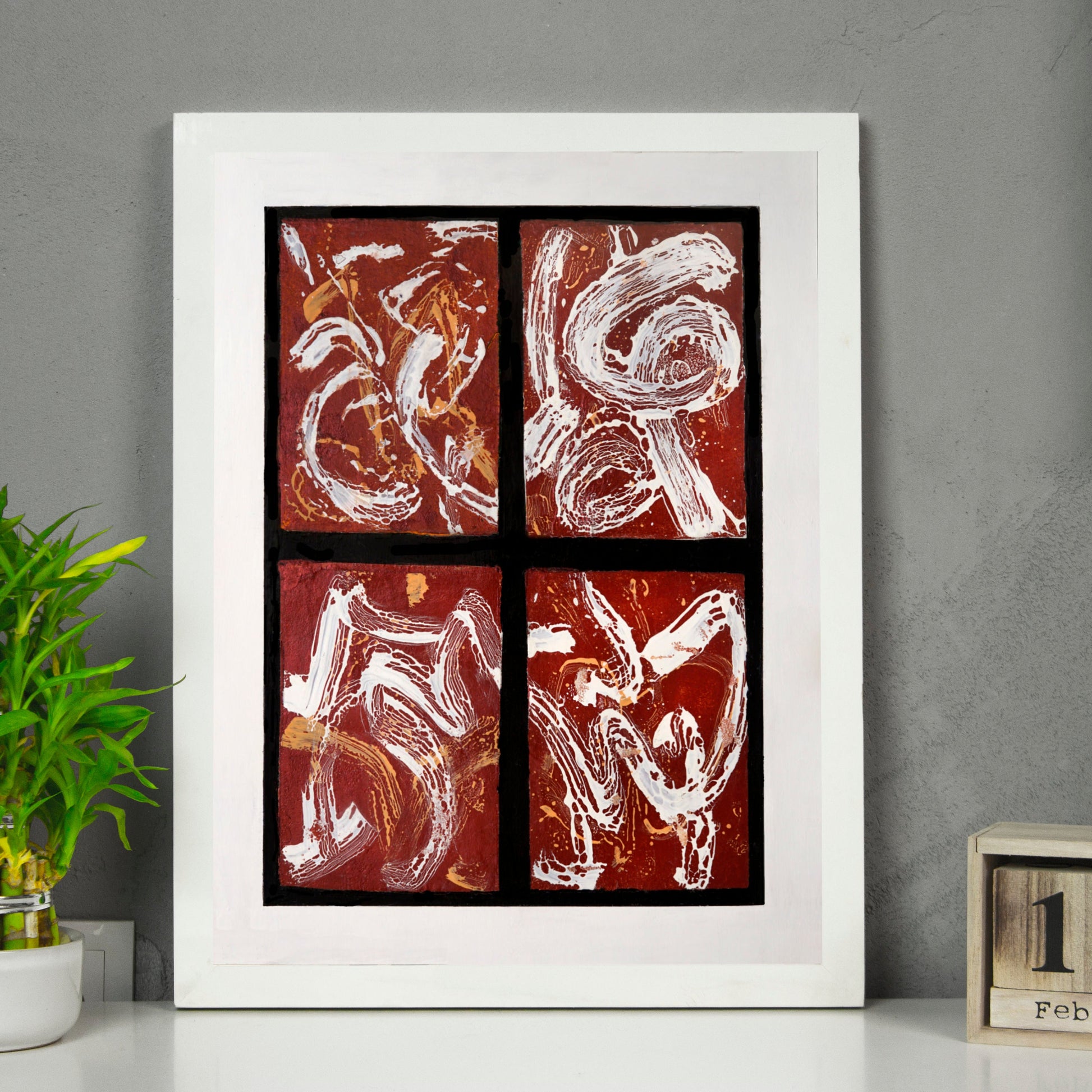 Framed abstract artwork of Chinese brush strokes on a wall with a plant and calendar below.