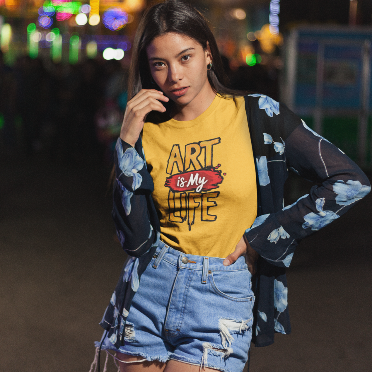 Woman wearing a yellow 'Art is My Love' t-shirt at a nighttime fair.