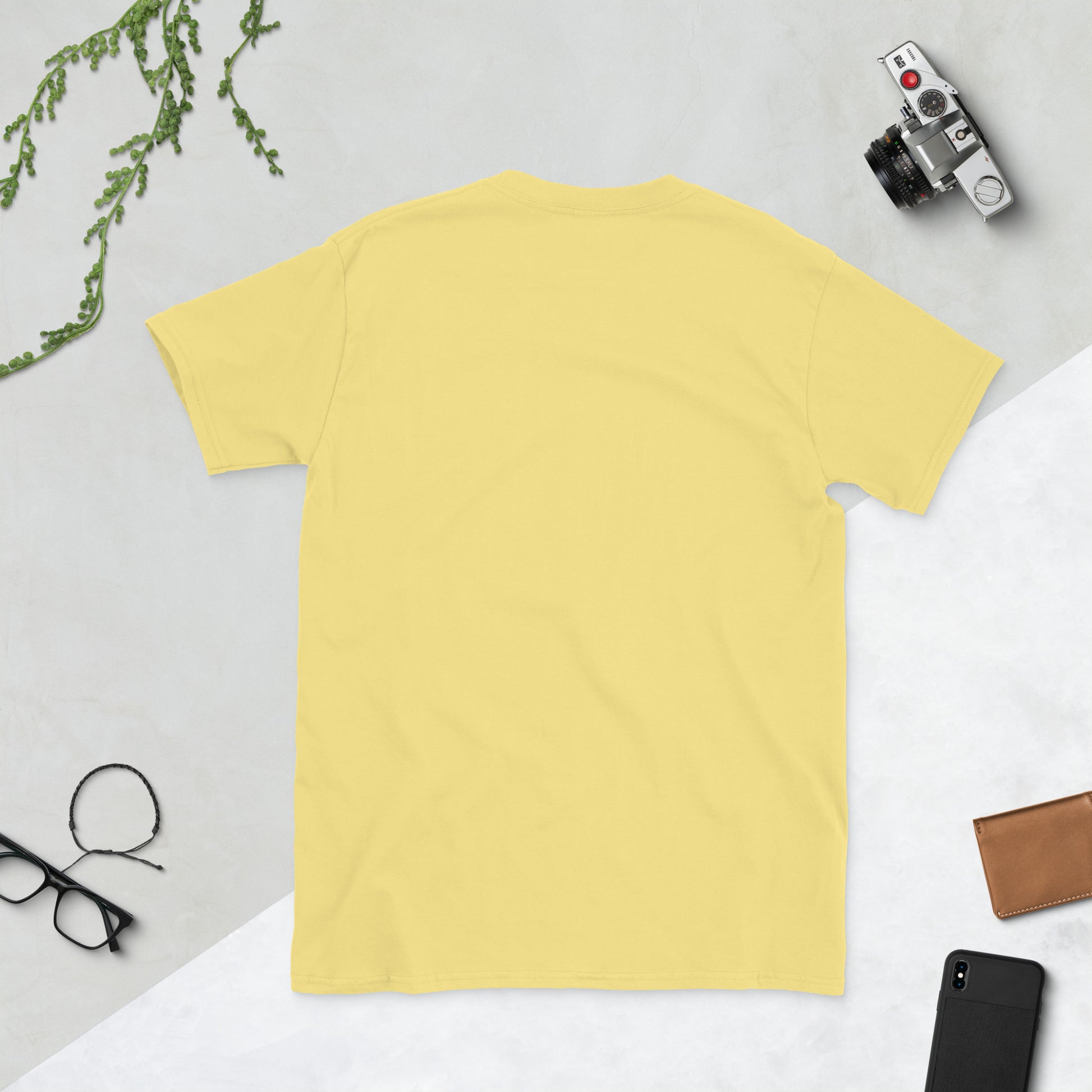 Yellow t-shirt on a light gray surface with a camera, phone, and glasses.