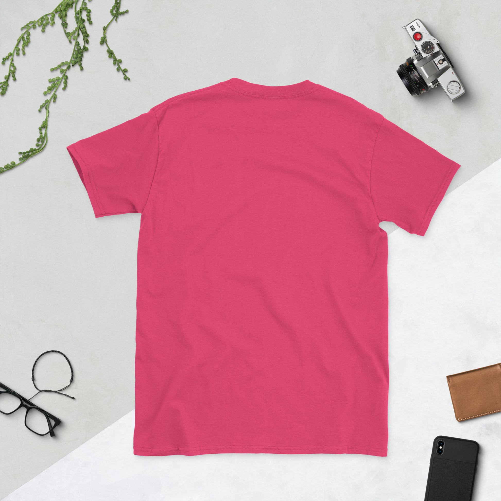 Pink t-shirt on a light gray surface with a camera, phone, and glasses.